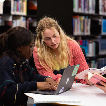 Two students sitting in the library, working together with laptops and notes