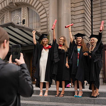 Four graduating students wearing dresses, gowns and hats holding their scrolls in the air while having their photo taken