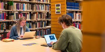 Two students sitting in library studying with laptops