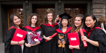 A group of students in graduation gowns holding flowers, scrolls and a teddy bear to celebrate